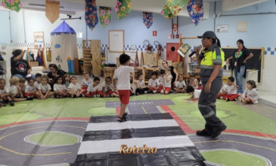Agente de trânsito com microfone conduz atividade educativa com crianças sentadas em tapete temático de trânsito no interior de escola em São José dos Campos, durante ação do programa Educamob no Maio Amarelo. Foto: Divulgação/Shopping Jardim Oriente