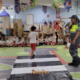 Agente de trânsito com microfone conduz atividade educativa com crianças sentadas em tapete temático de trânsito no interior de escola em São José dos Campos, durante ação do programa Educamob no Maio Amarelo. Foto: Divulgação/Shopping Jardim Oriente