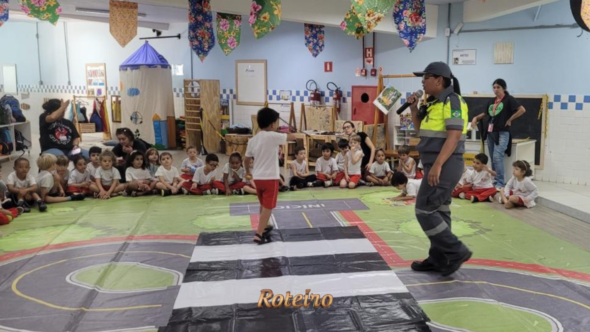 Agente de trânsito com microfone conduz atividade educativa com crianças sentadas em tapete temático de trânsito no interior de escola em São José dos Campos, durante ação do programa Educamob no Maio Amarelo. Foto: Divulgação/Shopping Jardim Oriente