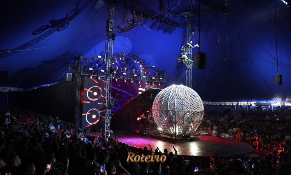 Vista interna do Mirage Circus com plateia lotada em ambiente de iluminação azul intensa. No centro da pista, uma grande gaiola esférica metálica iluminada. Ao fundo, um acrobata realiza manobra de moto em rampa elevada. Estrutura de treliças, telões de LED e efeitos de luz compõem o cenário grandioso do espetáculo.