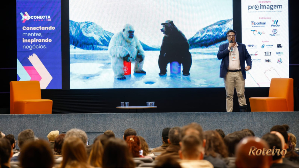 Palestrante do ProConecta Summit no auditório do PIT SJC, homem de blazer azul e microfone na mão, apresentando slide com criações em inteligência artificial projetadas em telão gigante. Plateia numerosa acompanha a apresentação. À direita, painel com patrocinadores e realizadores do evento.