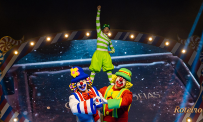 Três palhaços do Patati Patatá Circo Show em cena acrobática: dois palhaços coloridos em primeiro plano fazem formato de coração com as mãos enquanto sustentam um terceiro palhaço vestido de verde em pé sobre seus ombros, com o braço levantado em sinal de vitória. Ao fundo, cenário iluminado com moldura decorativa circense e telão estrelado.