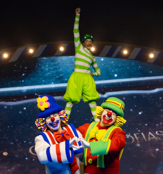 Três palhaços do Patati Patatá Circo Show em cena acrobática: dois palhaços coloridos em primeiro plano fazem formato de coração com as mãos enquanto sustentam um terceiro palhaço vestido de verde em pé sobre seus ombros, com o braço levantado em sinal de vitória. Ao fundo, cenário iluminado com moldura decorativa circense e telão estrelado.