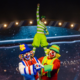 Três palhaços do Patati Patatá Circo Show em cena acrobática: dois palhaços coloridos em primeiro plano fazem formato de coração com as mãos enquanto sustentam um terceiro palhaço vestido de verde em pé sobre seus ombros, com o braço levantado em sinal de vitória. Ao fundo, cenário iluminado com moldura decorativa circense e telão estrelado.