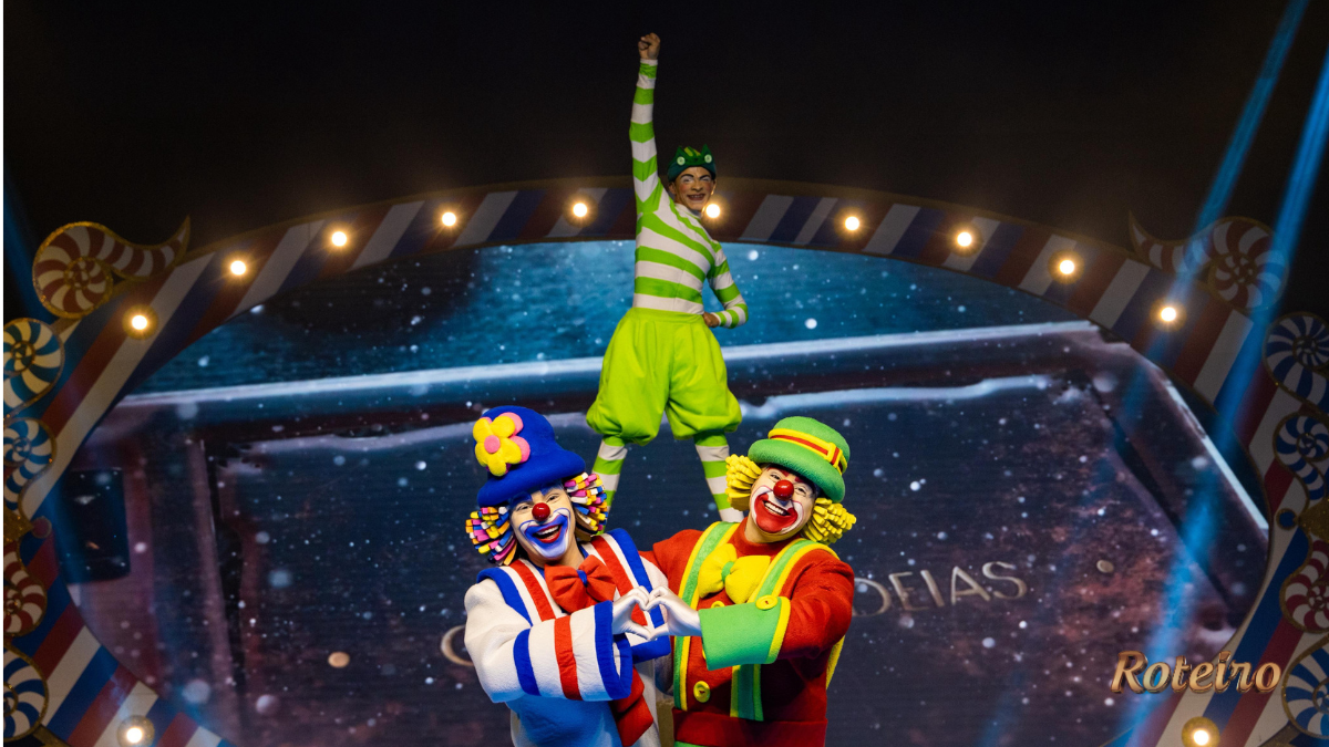 Três palhaços do Patati Patatá Circo Show em cena acrobática: dois palhaços coloridos em primeiro plano fazem formato de coração com as mãos enquanto sustentam um terceiro palhaço vestido de verde em pé sobre seus ombros, com o braço levantado em sinal de vitória. Ao fundo, cenário iluminado com moldura decorativa circense e telão estrelado.
