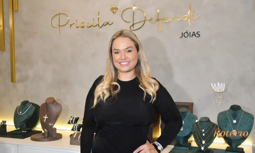 Empresária Priscila Defendi sorri em frente ao painel da marca Priscila Defendi Jóias, com expositores de colares, crucifixos e joias em ouro e prata ao redor, durante coquetel de apresentação da joalheria em São José dos Campos