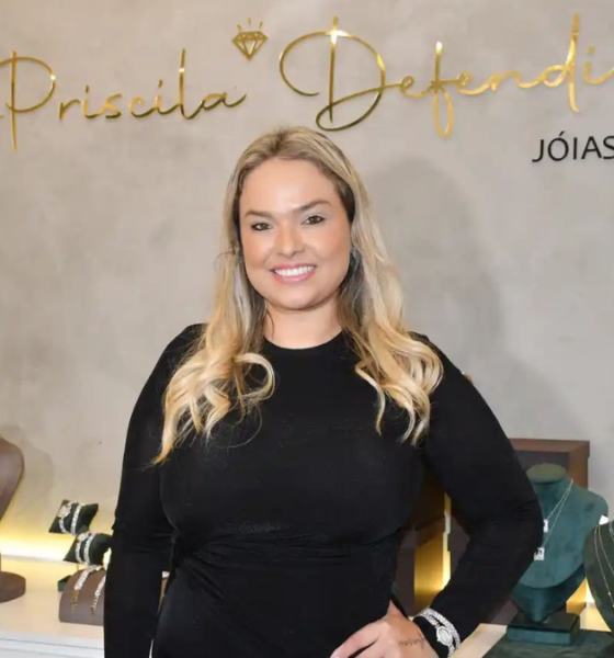 Empresária Priscila Defendi sorri em frente ao painel da marca Priscila Defendi Jóias, com expositores de colares, crucifixos e joias em ouro e prata ao redor, durante coquetel de apresentação da joalheria em São José dos Campos