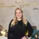 Empresária Priscila Defendi sorri em frente ao painel da marca Priscila Defendi Jóias, com expositores de colares, crucifixos e joias em ouro e prata ao redor, durante coquetel de apresentação da joalheria em São José dos Campos
