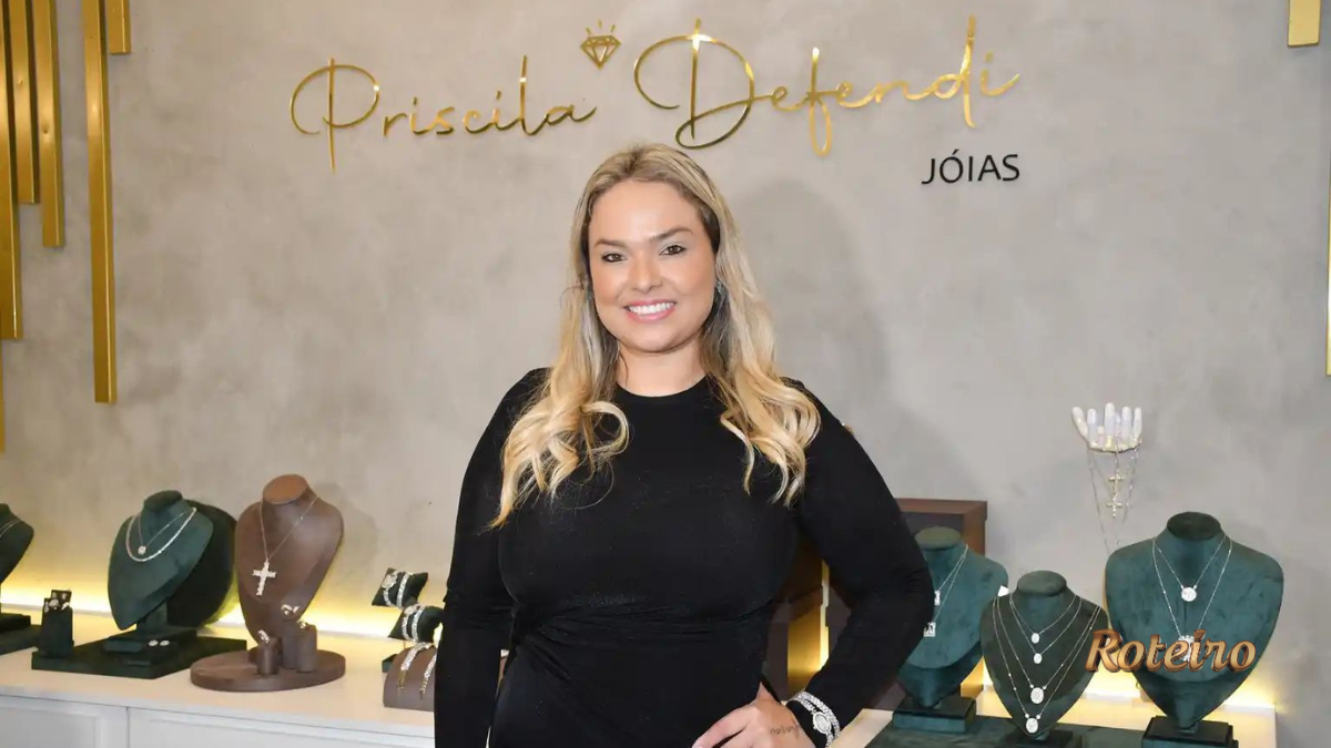 Empresária Priscila Defendi sorri em frente ao painel da marca Priscila Defendi Jóias, com expositores de colares, crucifixos e joias em ouro e prata ao redor, durante coquetel de apresentação da joalheria em São José dos Campos
