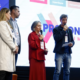 Quatro pessoas em pé no palco do ProConecta Summit 2025, no PIT São José dos Campos. Ao fundo, telão exibe o logo do evento e as trilhas: Audiovisual, Experiência, Comunicação e Design, e Empreendedorismo. Uma mulher de vestido vermelho e um homem de jaqueta preta seguram microfones durante apresentação.