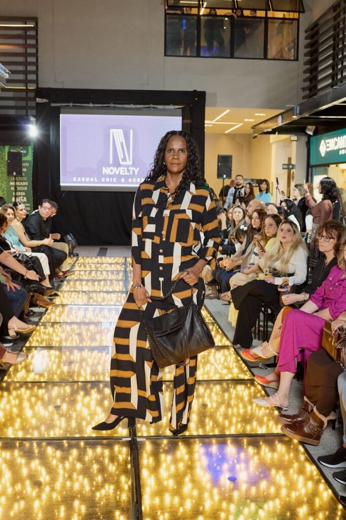 Modelo apresenta look da loja Novelty Casual Chic na passarela iluminada do desfile de outono/inverno do Pátio Villa Branca em Jacareí, vestindo conjunto estampado preto, bege e dourado com bolsa preta, diante de público numeroso