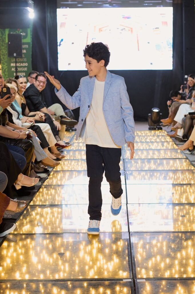 Criança desfila na passarela iluminada do desfile especial de outono/inverno do Pátio Villa Branca em Jacareí, vestindo blazer azul claro, calça preta e tênis bicolor, com público ao redor aplaudindo