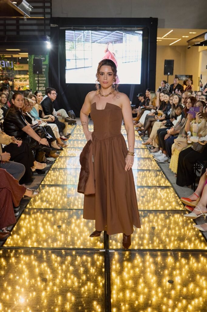Modelo desfila na passarela iluminada do Pátio Villa Branca em Jacareí com vestido midi marrom tomara que caia e casaco na mão, durante o desfile especial de outono/inverno, com plateia lotada ao fundo