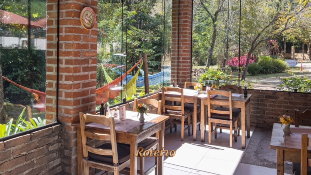 Salão envidraçado do restaurante Donna Pinha em Santo Antônio do Pinhal, com mesas e cadeiras de madeira rústica, paredes de tijolo à vista e vista para jardim com redes coloridas ao fundo. Foto: Douglas Fagundes
