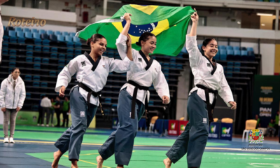 Três atletas da equipe Liga Vale de São José dos Campos com faixa preta celebram vitória segurando a bandeira do Brasil na Arena Carioca, no Rio de Janeiro, durante o Pan-Americano de Taekwondo 2026