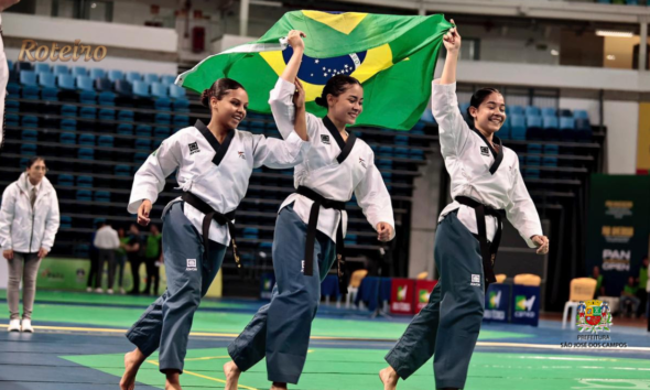 Três atletas da equipe Liga Vale de São José dos Campos com faixa preta celebram vitória segurando a bandeira do Brasil na Arena Carioca, no Rio de Janeiro, durante o Pan-Americano de Taekwondo 2026