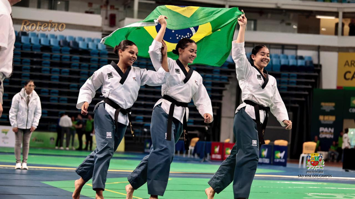 Três atletas da equipe Liga Vale de São José dos Campos com faixa preta celebram vitória segurando a bandeira do Brasil na Arena Carioca, no Rio de Janeiro, durante o Pan-Americano de Taekwondo 2026