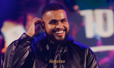 Cantor Yan sorridente no palco, vestindo jaqueta de couro preta e segurando microfone, com iluminação roxa e cenário colorido ao fundo durante show. Foto: Divulgação