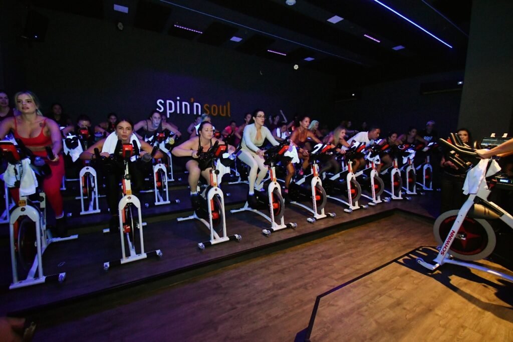 Alunos pedalando em aula de indoor bike com bicicletas Schwinn na sala escura da Spin'n Soul SJC, com o logo da marca iluminado ao fundo. Foto: Divulgação