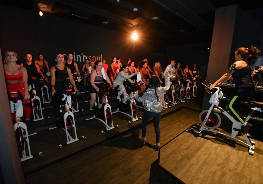 Instrutora conduz aula de indoor bike na Spin'n Soul SJC com turma completa ao fundo e iluminação de palco. Foto: Divulgação