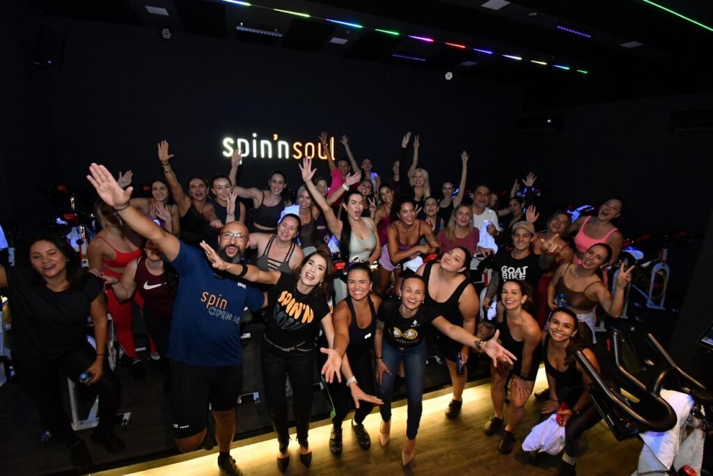 Grupo de alunos e equipe da Spin'n Soul SJC celebra com os braços erguidos após aula inaugural, em frente ao logo iluminado da marca na parede da sala de spinning. Foto: Divulgação