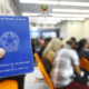 Pessoa segurando Carteira de Trabalho e Previdência Social em sala de espera do PAT de São José dos Campos com candidatos ao fundo
