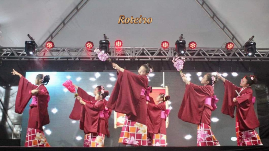 Grupo de dançarinas com quimono vinho e laranja realiza a dança da cerejeira no palco iluminado do Vale Ninhon Matsuri, segurando flores cor-de-rosa. Foto: Divulgação