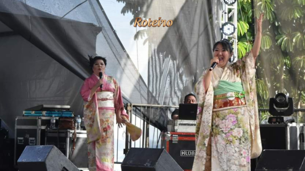 Duas cantoras vestindo kimono colorido se apresentam no palco do Vale Ninhon Matsuri com microfone, em show de música típica japonesa ao ar livre. Foto: Divulgação