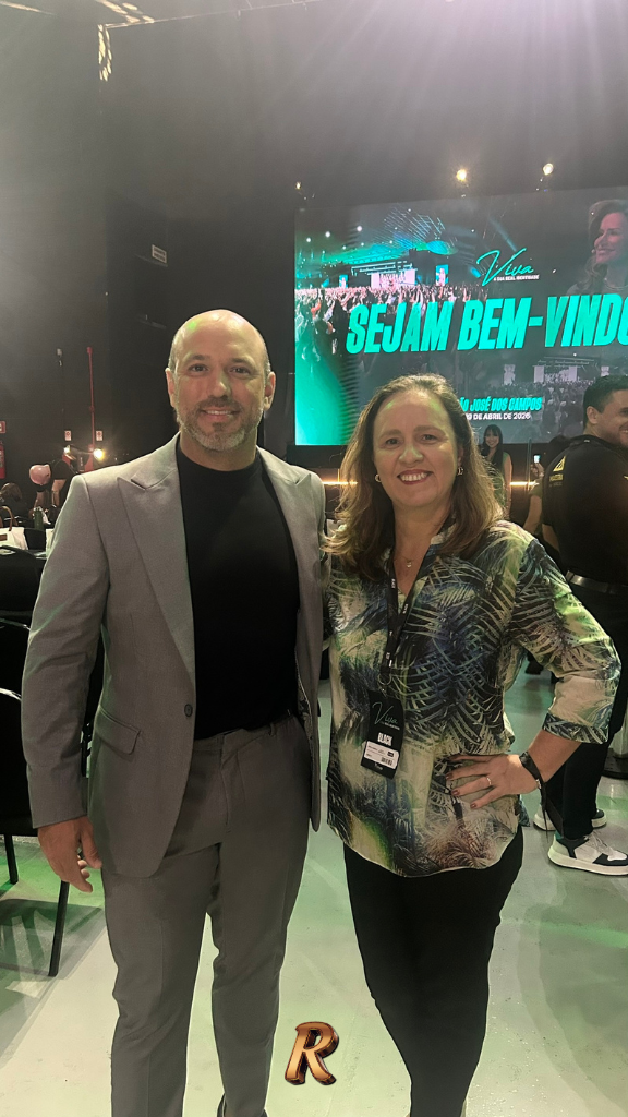 Vinícius Carlo e Patricia Morello posam juntos no evento Viva, em São José dos Campos, em abril de 2026. Vinícius veste terno cinza com camiseta preta; Patricia, blusa estampada. Ao fundo, telão com a mensagem "Sejam Bem-Vindos" e a data do evento.