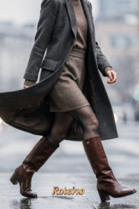 Mulher de bota cano longo marrom e sobretudo cinza atravessando rua — calçados tendência moda inverno 2026 SJC