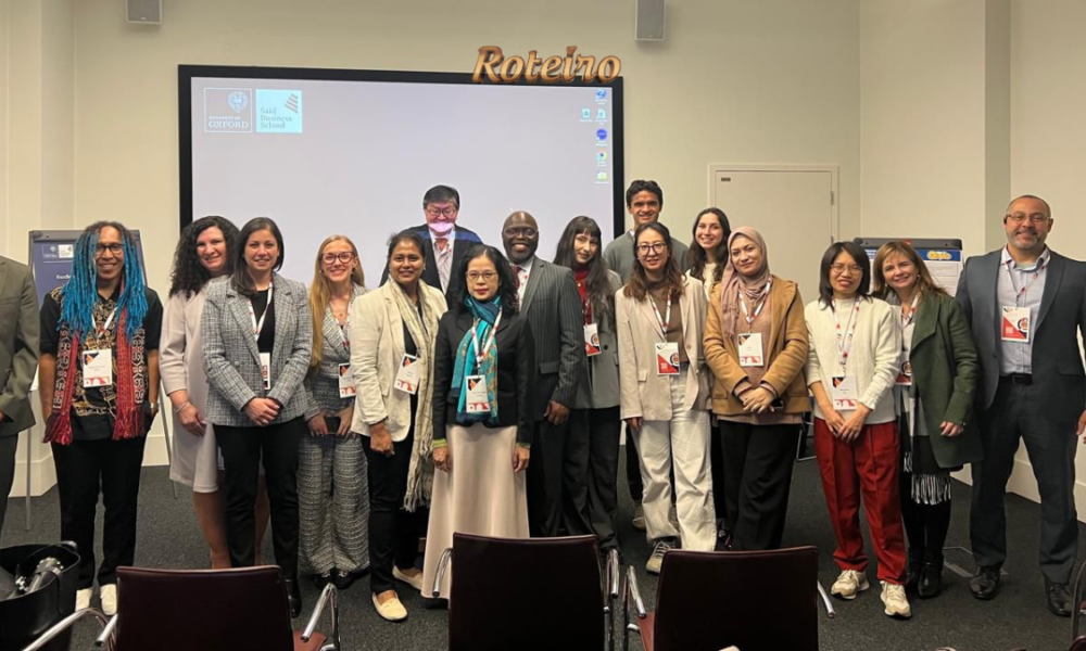 Grupo de cerca de 18 participantes do congresso internacional posa para foto em sala da Saïd Business School da University of Oxford. Ao fundo, projetor exibe os logos da universidade. Logo do Portal RoteiroSJC no topo da imagem.