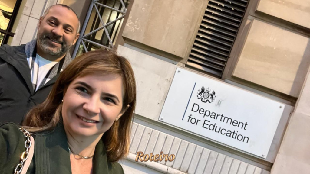 Casal posa sorridente em frente à placa oficial do Department for Education do governo britânico, em Londres. Logo do Portal RoteiroSJC na imagem.