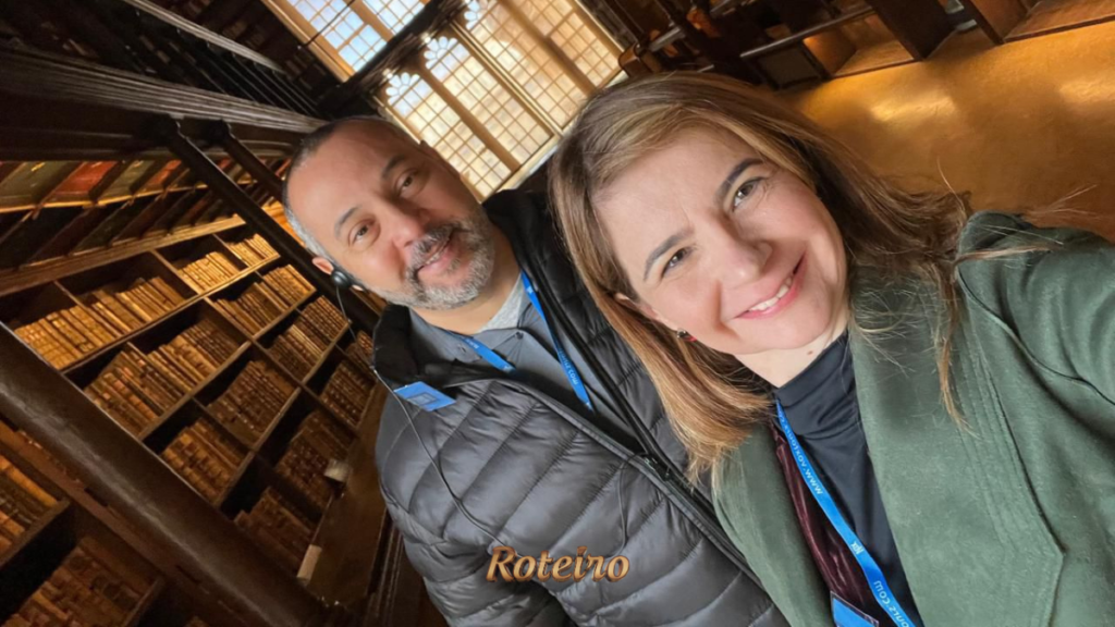 Casal tira selfie no interior de biblioteca histórica da University of Oxford, com prateleiras repletas de livros antigos ao fundo e iluminação quente. Ambos usam crachás de identificação do congresso. Logo do Portal RoteiroSJC na imagem.