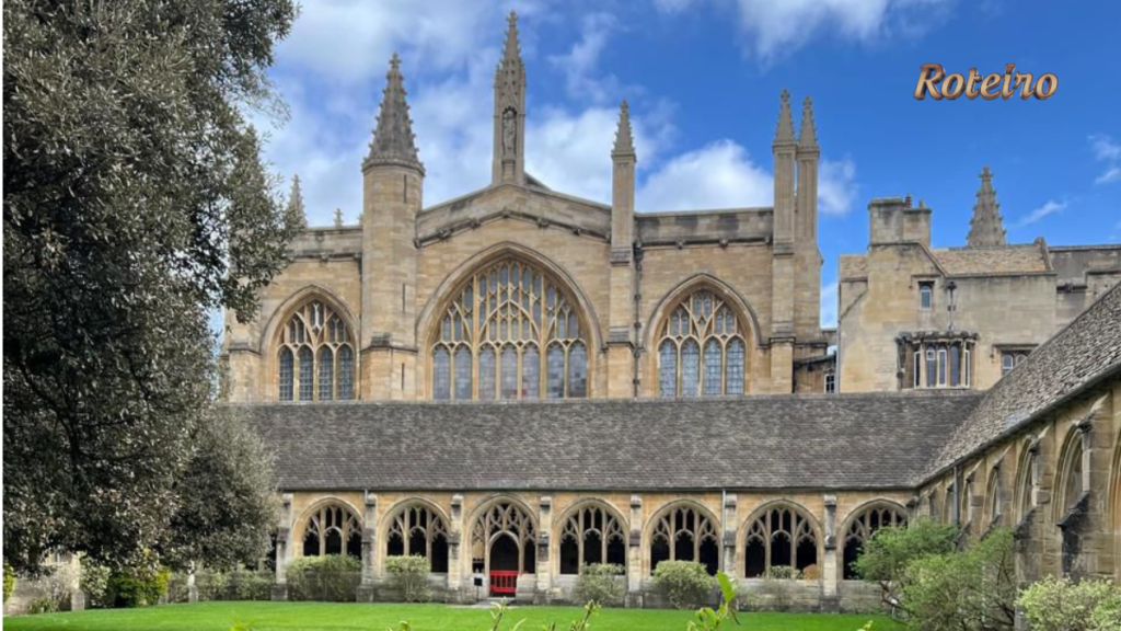 Fachada gótica da University of Oxford com claustro em arcos medievais, gramado verde e torres pontiagudas em pedra dourada sob céu azul com nuvens. Logo do Portal RoteiroSJC no canto superior direito.