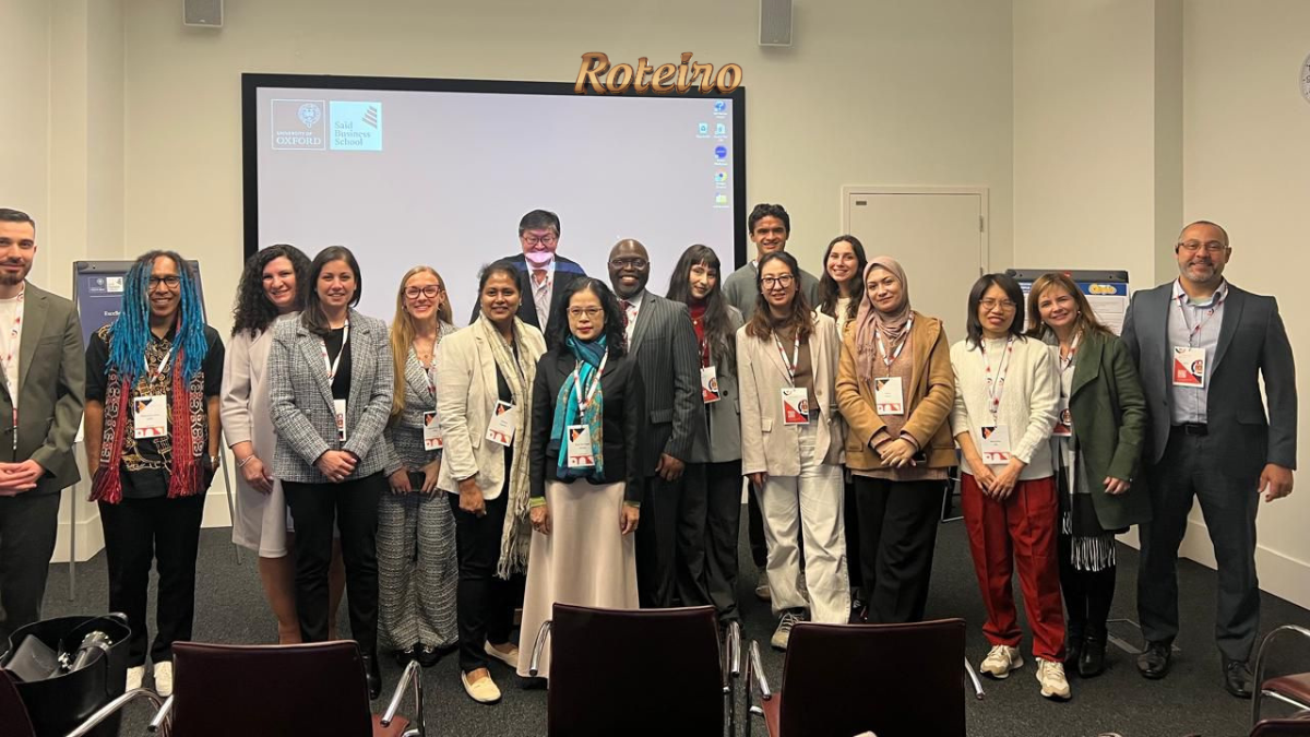 Grupo de cerca de 18 participantes do congresso internacional posa para foto em sala da Saïd Business School da University of Oxford. Ao fundo, projetor exibe os logos da universidade. Logo do Portal RoteiroSJC no topo da imagem.