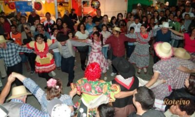 Festas Juninas SJC 2026. Pátio lotado, pessoas a carater dançando