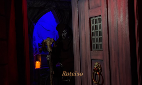 Halloween Castelo do Terror: personagem encapuzado com máscara de caveira e cajado surge atrás de porta de madeira no percurso da atração